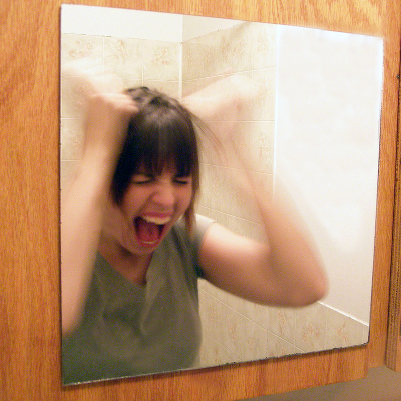 A woman stands in front of a mirror screaming and pulling her own hair. A woman stands in front of a mirror screaming and pulling her own hair.