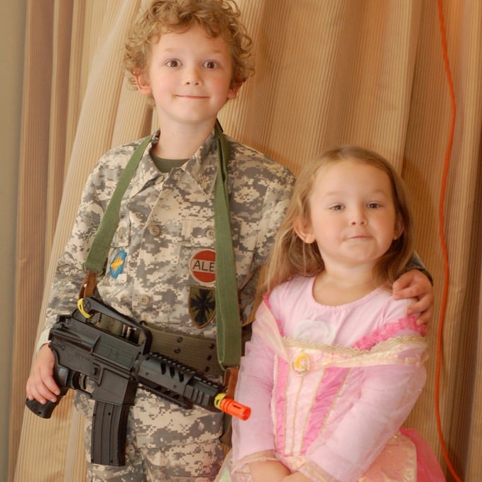 A brother and sister stand side by side. He is dressed in a camouflage military uniform and is holding a toy gun. She is dressed in a pretty pink princess dress. A brother and sister stand side by side. He is dressed in a camouflage military uniform and is holding a toy gun. She is dressed in a pretty pink princess dress.