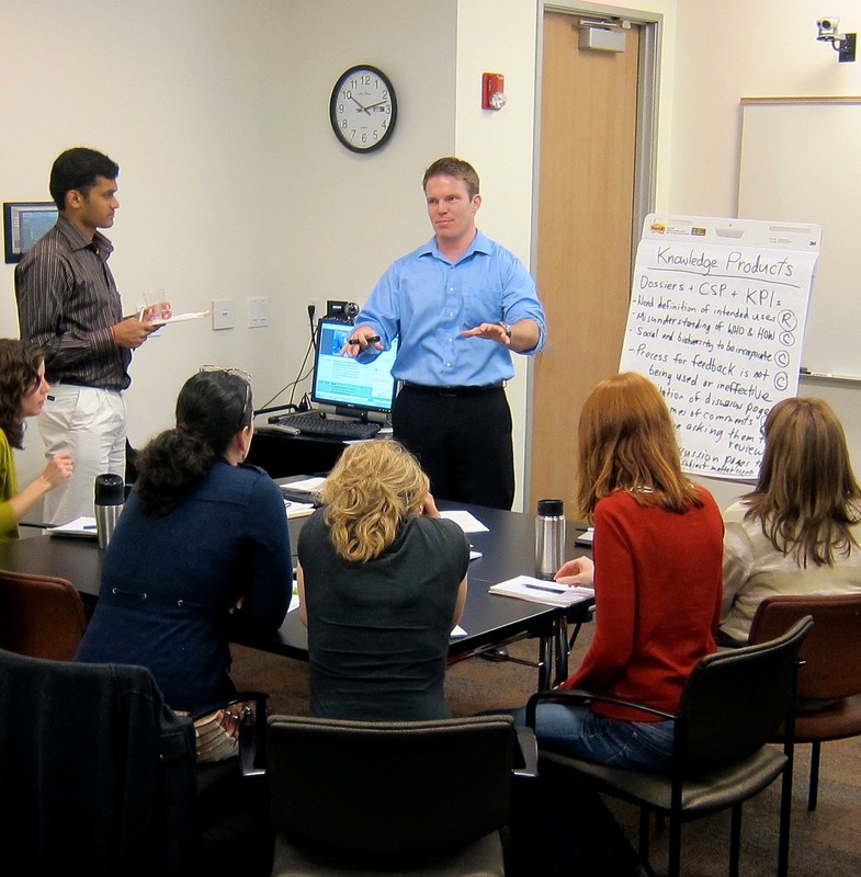 A group of workers are gathered around a conference table as a colleague makes a presentation.