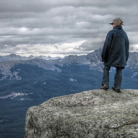 A boy stands at the edge of a cliff and looks out at a valley below and snow covered peaks in the distance. A boy stands at the edge of a cliff and looks out at a valley below and snow covered peaks in the distance.