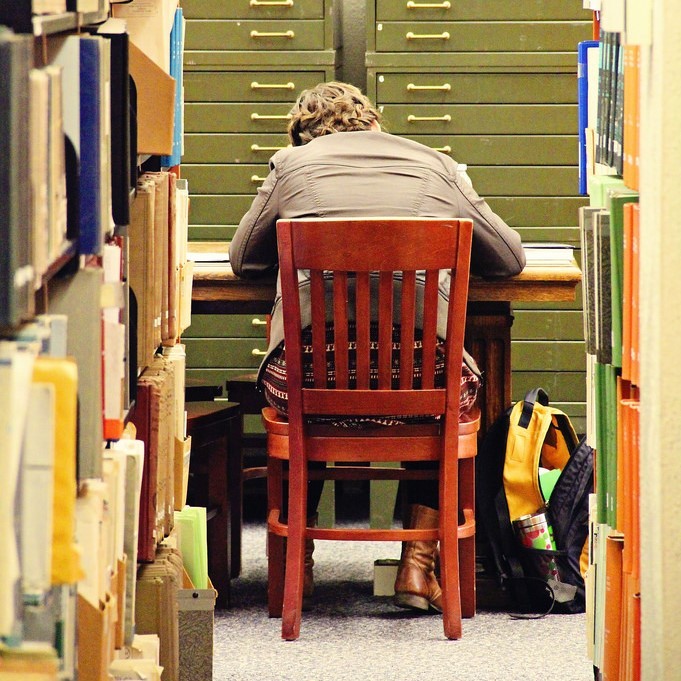 A student is hunched over a desk in the library. A student is hunched over a desk in the library.