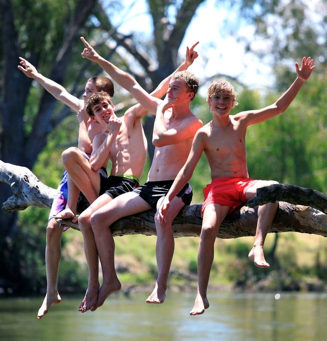 A group of teen aged boys in swimming trunks sit together on a tree branch above a pond as they smile and show off for the camera. A group of teen aged boys in swimming trunks sit together on a tree branch above a pond as they smile and show off for the camera.