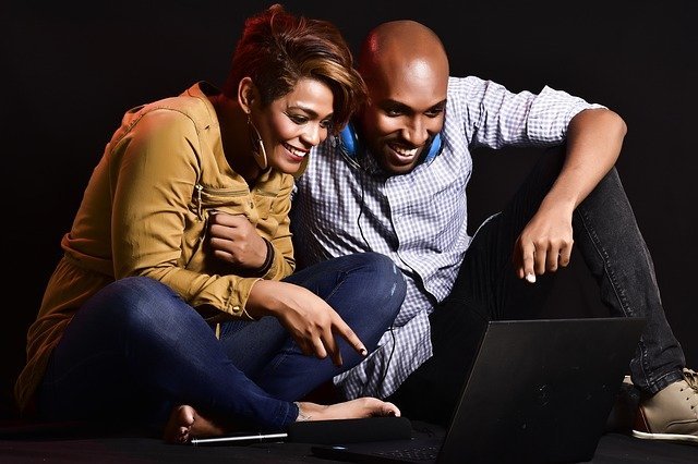A young couple smiling and watching something on their laptop screen, the woman is pointing something out to the man.